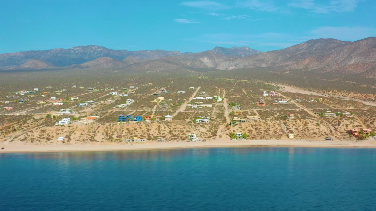 Wide drone frame over turquoise water toward El Sargento La Ventana, Mexico, with a sandy beach, scattered coastal buildings, dry roads, and mountains in the distance, no visible people.