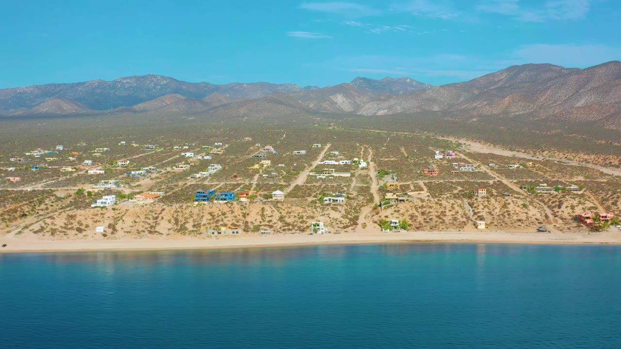 Aerial frame of El Sargento La Ventana coastline with calm blue-green water in front, a narrow sand strip, low buildings and roads behind it, and hazy mountains under clear sky, no visible people.