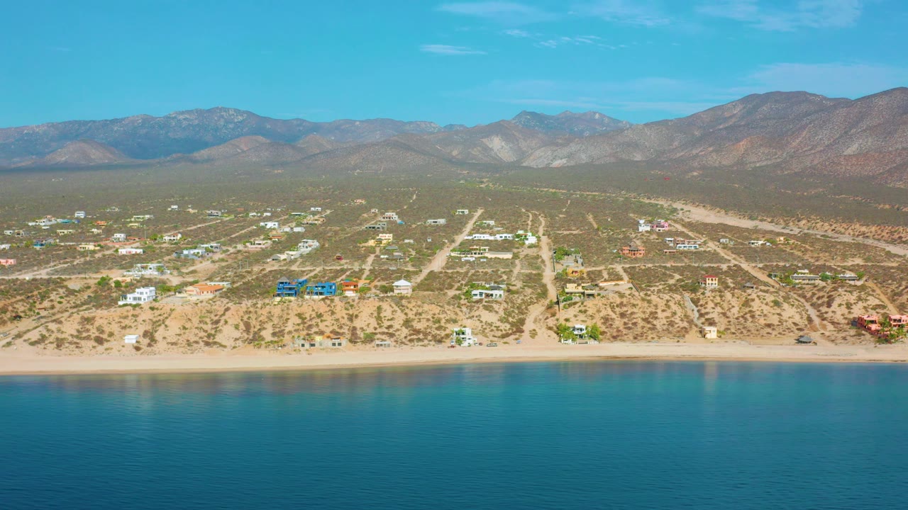 Aerial coastline frame with turquoise foreground water and dry inland roads below mountains.