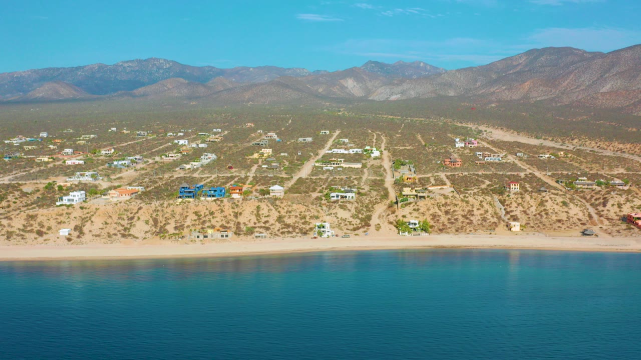 At 0:00, a wide aerial frame over El Sargento La Ventana shows turquoise water in front, a sandy beach and scattered buildings inland, and brown mountains under a clear sky, with no visible people.