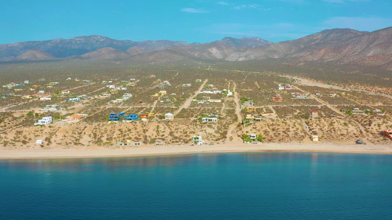 At 0:01, the aerial view holds over El Sargento La Ventana with blue water across the foreground, pale dirt roads and homes beyond the beach, and a long mountain ridge in back, with no visible people.