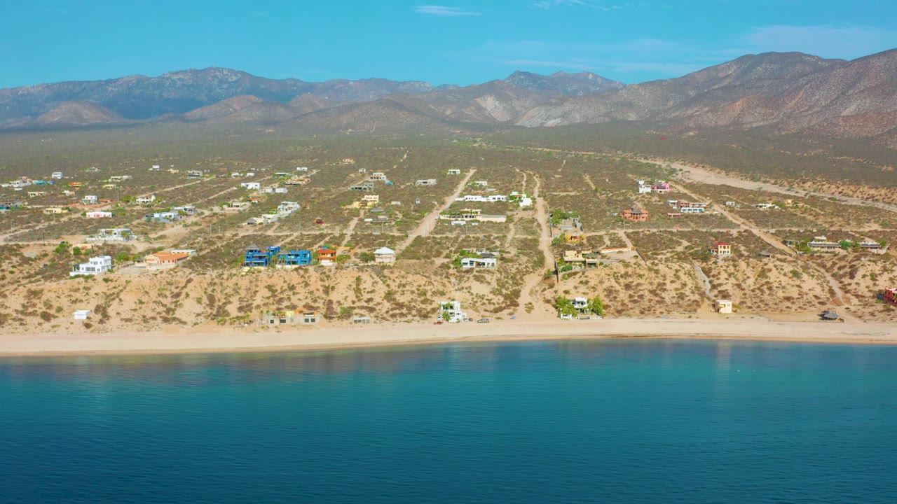 At 0:02, El Sargento La Ventana appears from offshore with calm sea in the foreground, a narrow sand strip, scattered houses on scrubby terrain, and mountains stretching across the horizon, with no visible people.