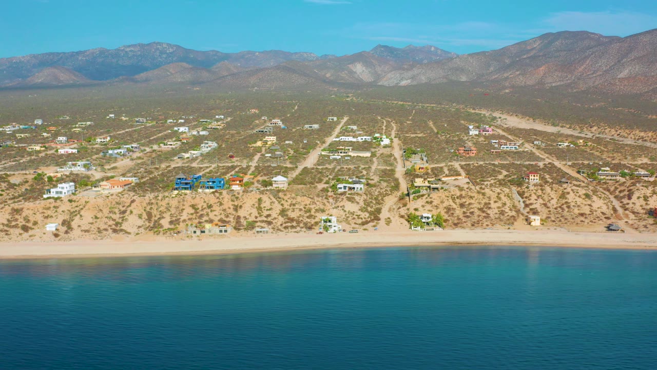 Aerial frame of beach, coastal houses, dirt road network, open scrubland, and mountains.