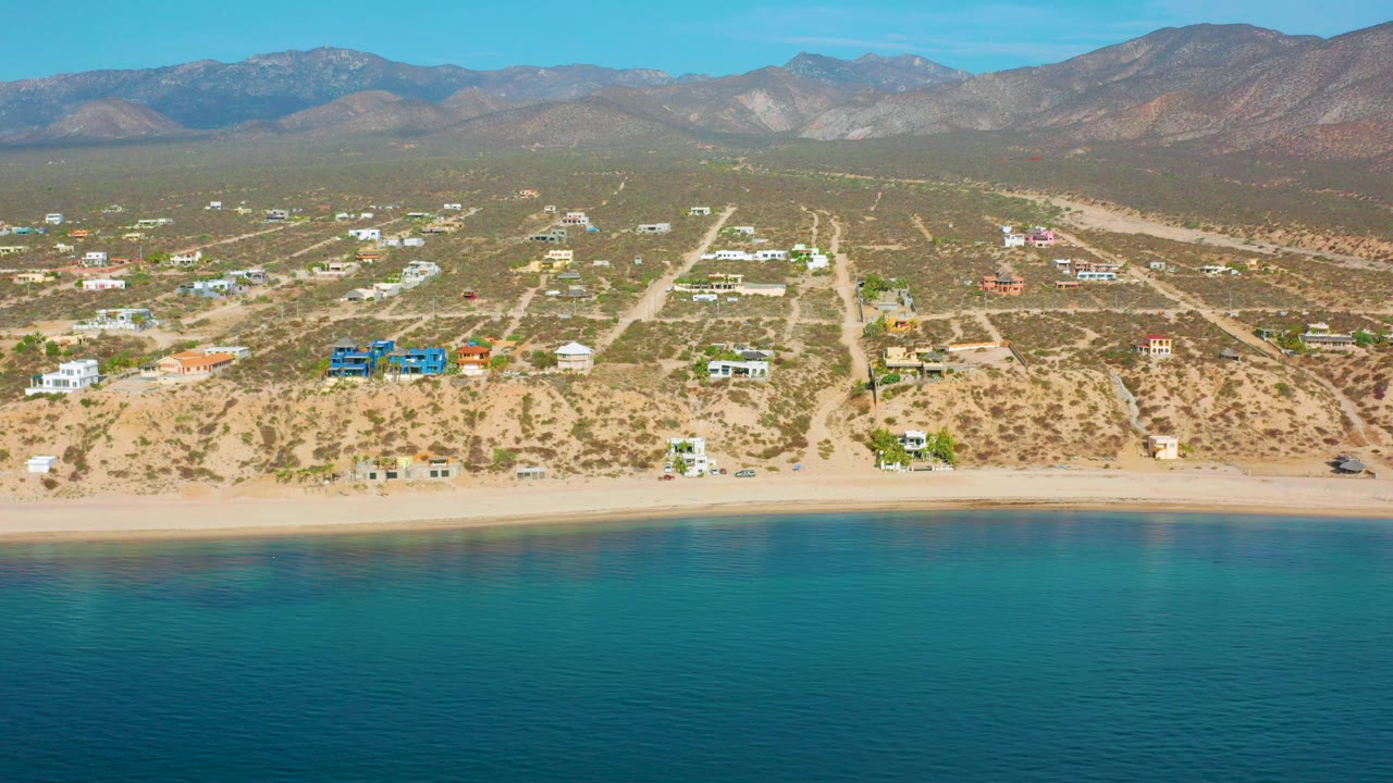 Opening aerial frame over El Sargento La Ventana, Mexico, with turquoise water in front, a narrow sand beach, scattered homes and dirt roads inland, and no visible people.