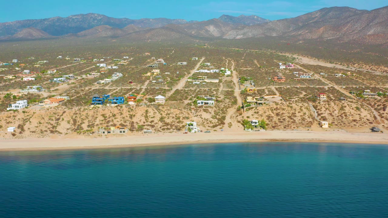 Wide drone view of beach, sparse houses, desert lots, and mountain backdrop.
