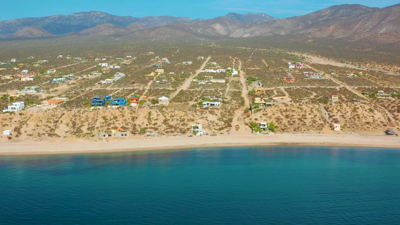 Aerial frame over El Sargento La Ventana, Mexico, with calm blue-green water at the bottom, a light sand strip, colorful homes inland, and no visible people.