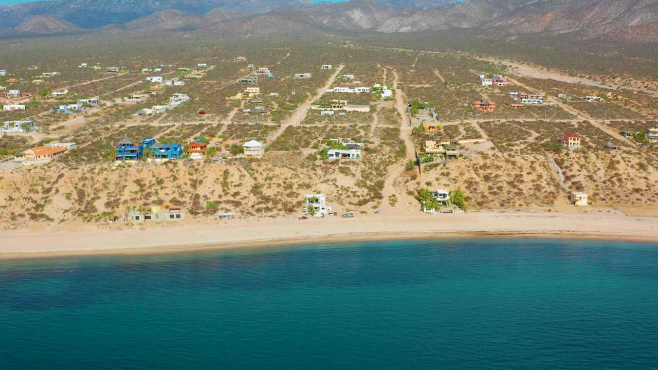 Opening aerial frame over El Sargento La Ventana, Mexico with turquoise ocean in front, a pale beach, scattered houses across desert lots, distant mountains, and no visible people.