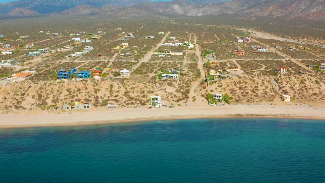 Wide drone view of beach houses behind a sandy shore with blue-green water below.