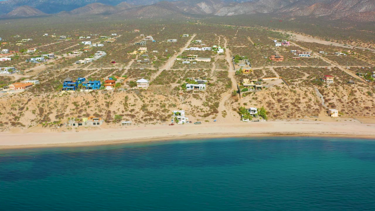 Aerial frame over El Sargento La Ventana, Mexico with teal water filling the lower frame, a beige beach line, small homes and roads across scrubland, hills beyond, and no visible people.