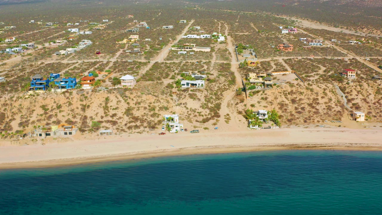 Aerial frame over El Sargento La Ventana, Mexico, with turquoise ocean foreground, pale beach, scattered homes, dirt roads, desert scrub, and no visible people.