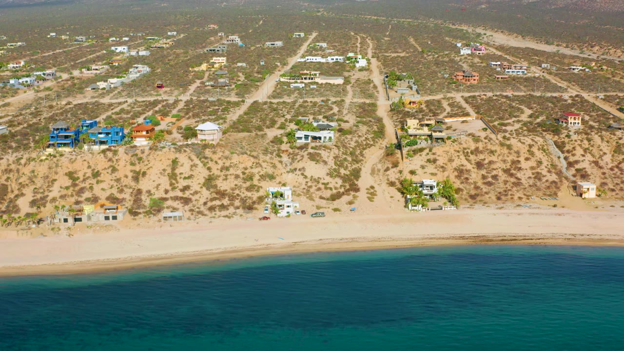 Aerial view in El Sargento La Ventana, Mexico, with the beach and water across the lower frame, scattered residences inland, dry lots, dirt access roads, and no visible people.