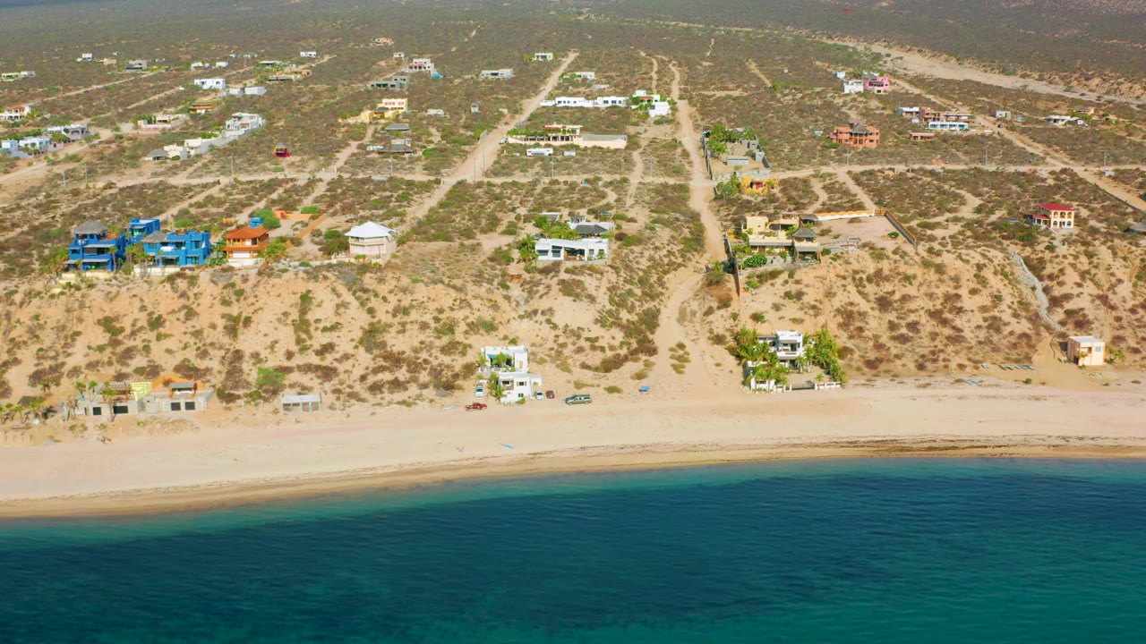 Drone aerial over El Sargento La Ventana, Mexico, showing blue beach-side buildings, nearby white and orange homes, sandy shore, green-blue water, scrubby terrain, and no visible people.