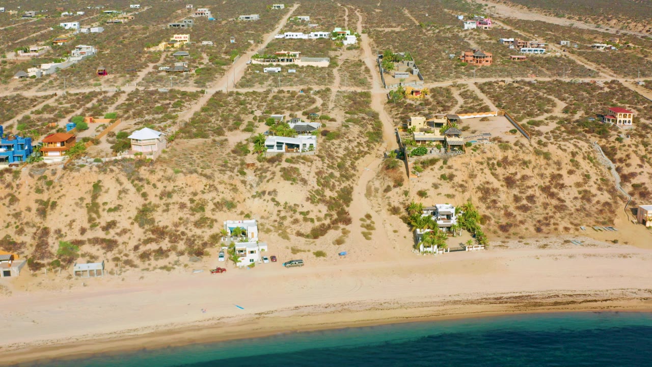 High aerial frame over El Sargento La Ventana, Mexico, showing scattered beach homes above a pale sandy shoreline and calm blue-green water, with no visible people.