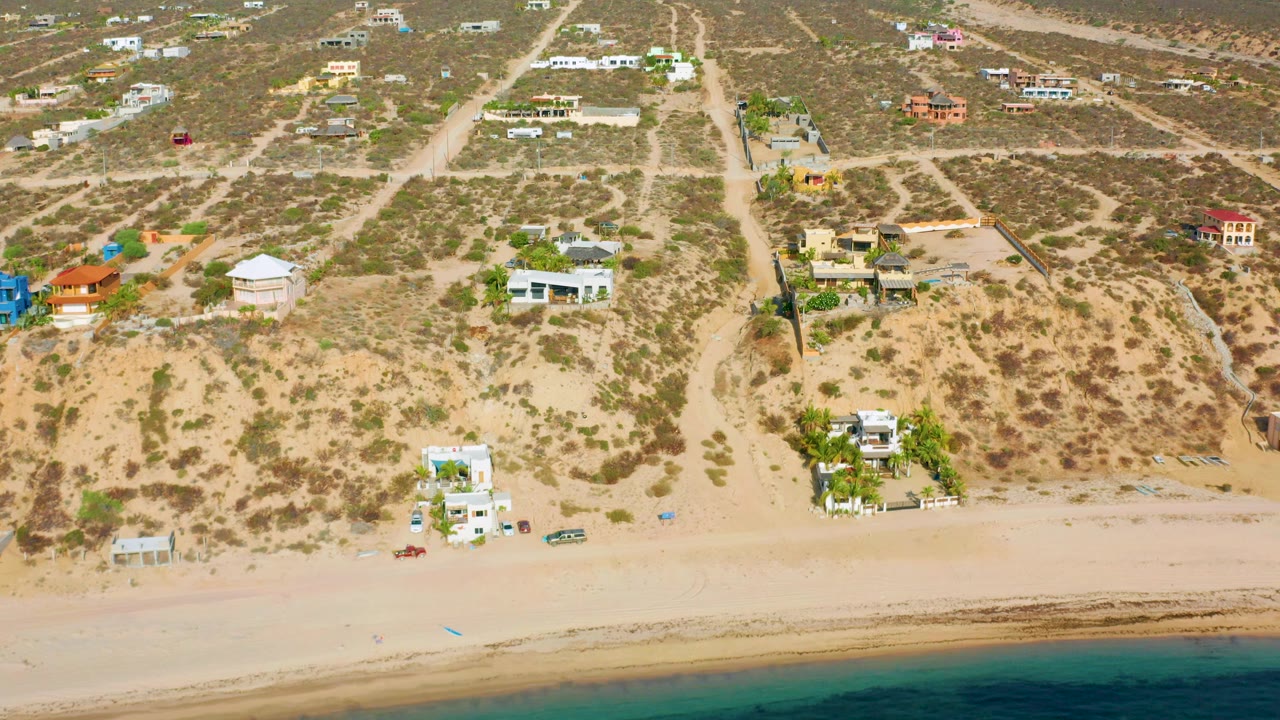 Aerial frame from El Sargento La Ventana, Mexico, showing open sandy beach, a small white home by the shore, inland dirt roads, dry brush, and no visible people.