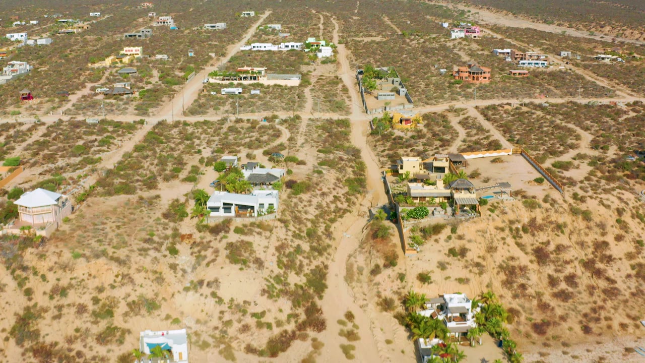 High oblique view in El Sargento La Ventana with dirt roads crossing scrubby lots, low homes on both sides, hills beyond, and no visible people.