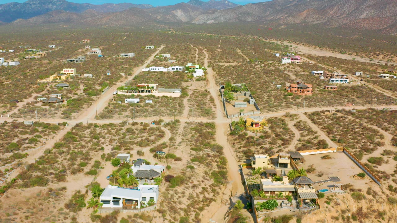 Aerial view over El Sargento La Ventana, Mexico, showing dirt roads, scattered desert buildings, scrubland, distant mountains, and no visible people.