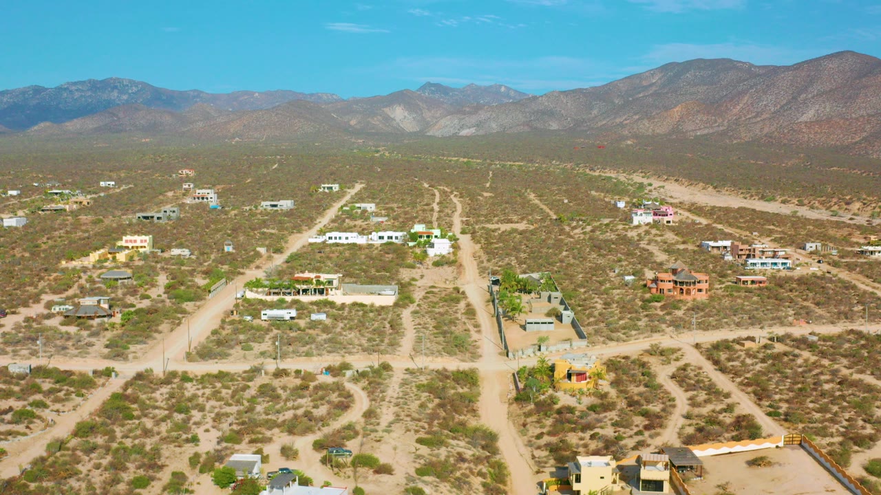 Opening aerial frame over El Sargento La Ventana, Mexico, showing intersecting dirt roads, low desert buildings, scrubland, distant mountains, and no visible people.