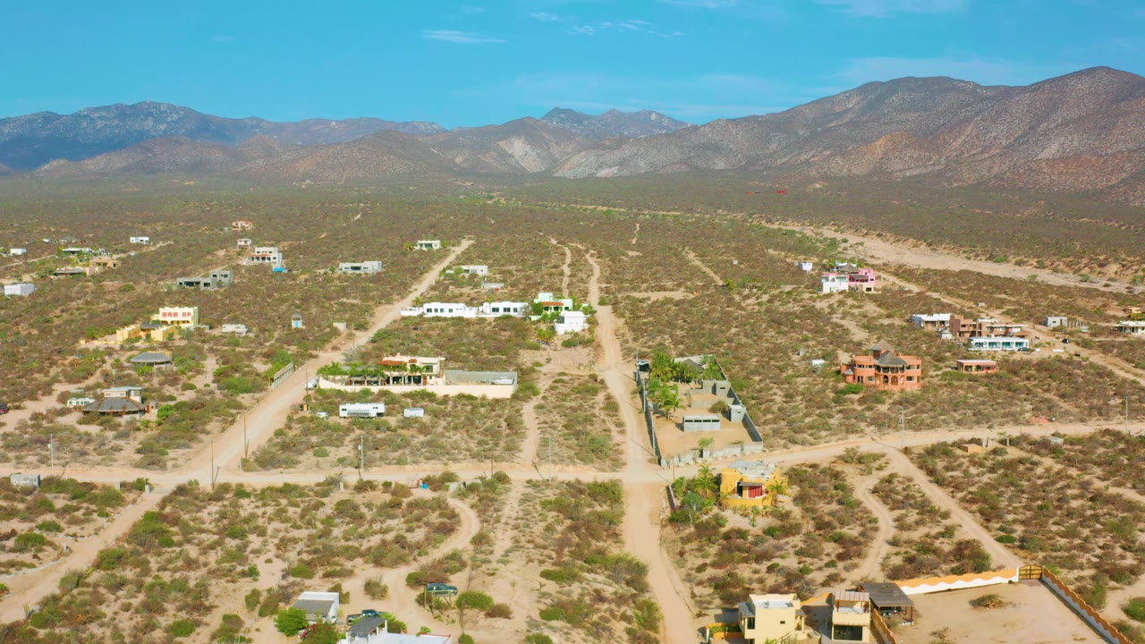 A second aerial view of El Sargento La Ventana, Mexico, with a central dirt road crossing scrubby lots, small buildings across the midground, mountain ridges beyond, and no visible people.