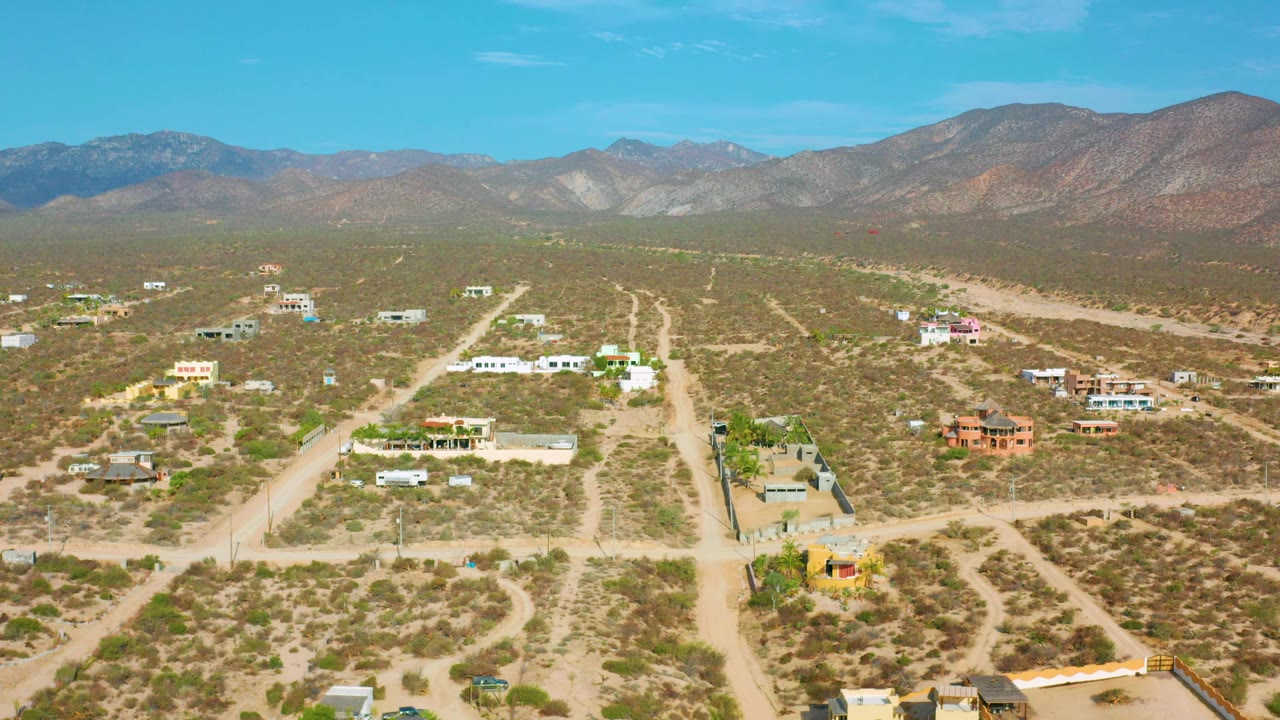 Aerial frame over El Sargento La Ventana, Mexico, with sandy roads leading through dry brush, several low structures in the midground, mountains spanning the background, and no visible people.