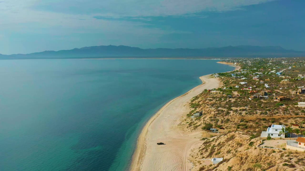 Aerial frame from El Sargento La Ventana, Mexico, showing the curved sand beach slightly farther along the coast, blue-green water on the left, homes and dirt roads on the right, mountains on the horizon, and no visible people.