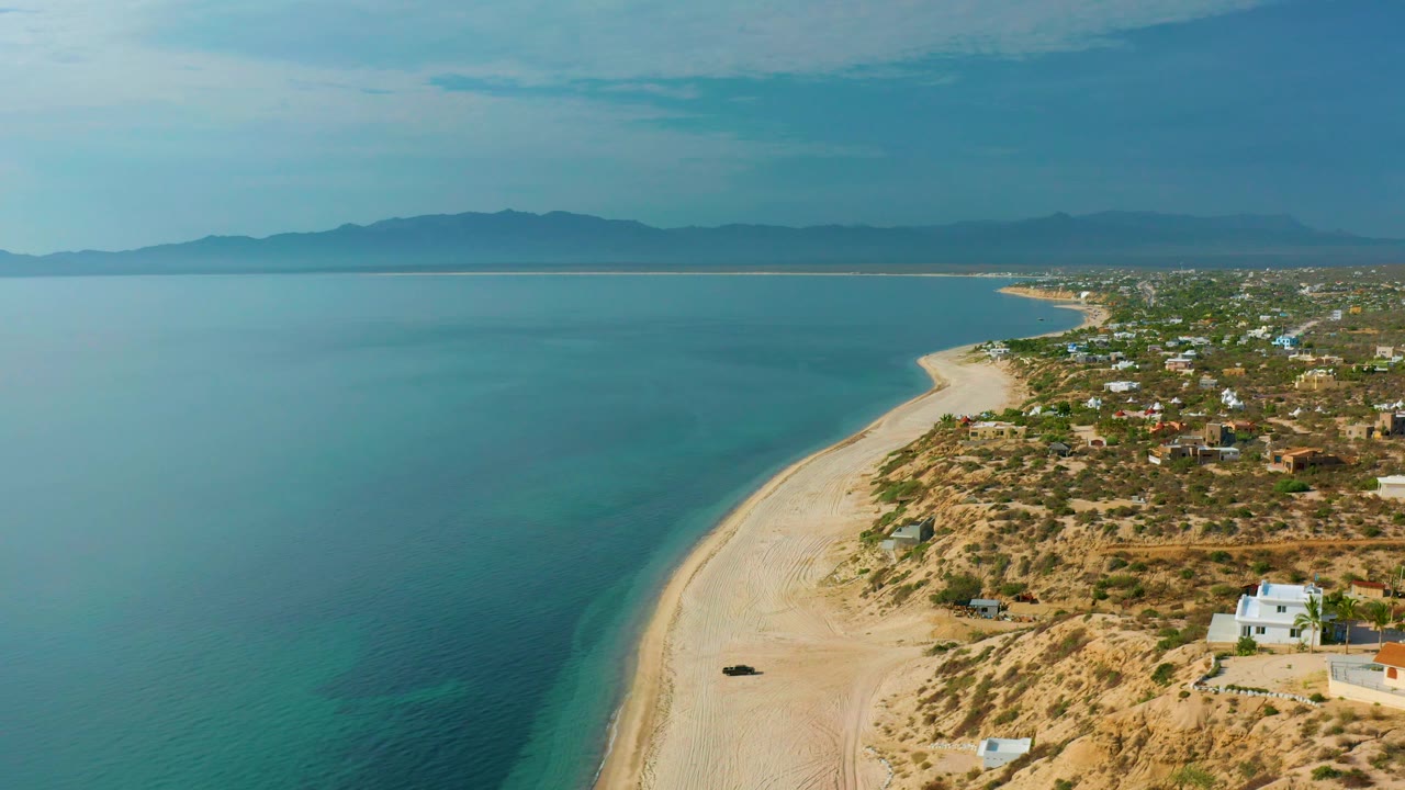 Wide aerial shoreline with calm water, sandy beach, desert terrain, and small homes.