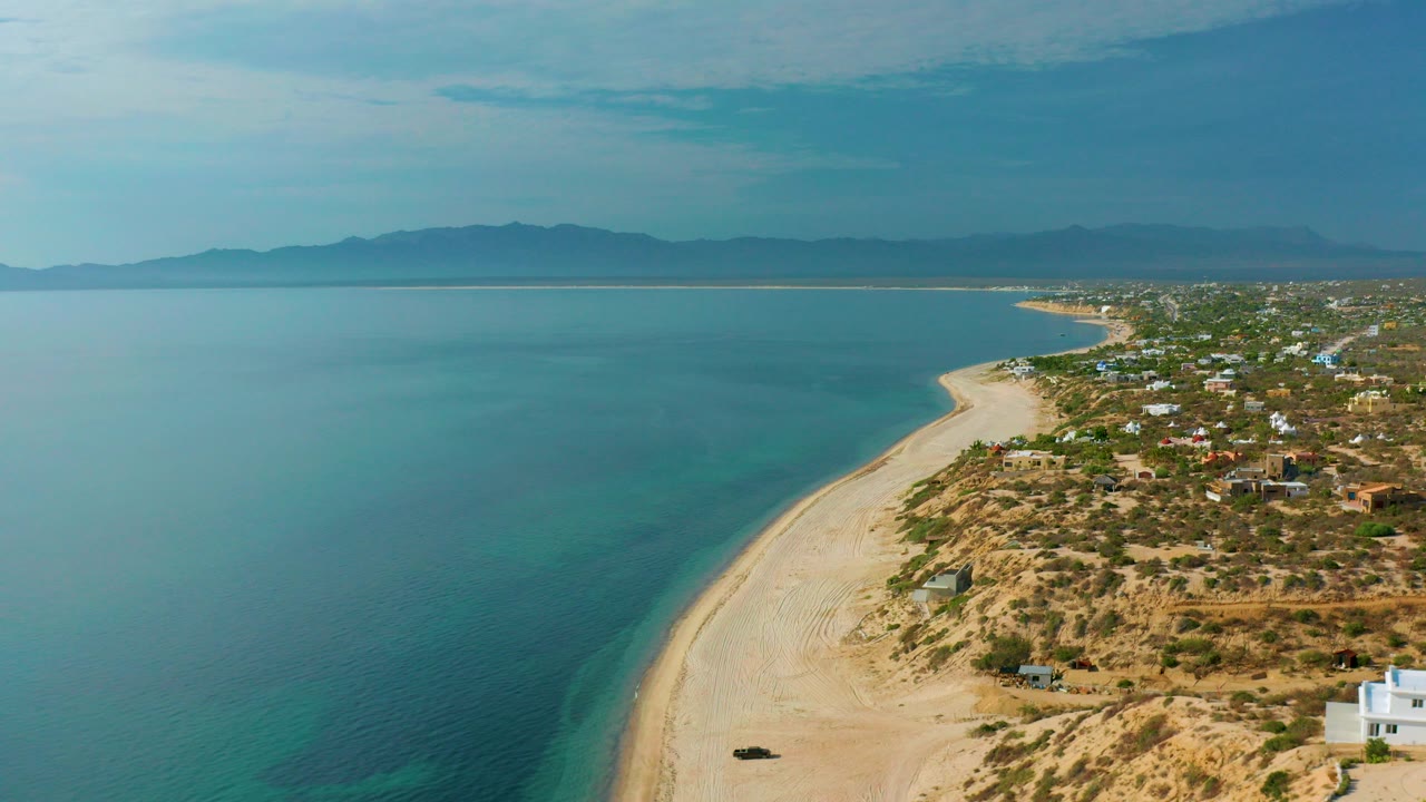 Opening aerial frame over El Sargento La Ventana, Mexico, with turquoise water at left, a pale beach curving along shore, scattered coastal buildings at right, hazy mountains beyond, and no visible people.