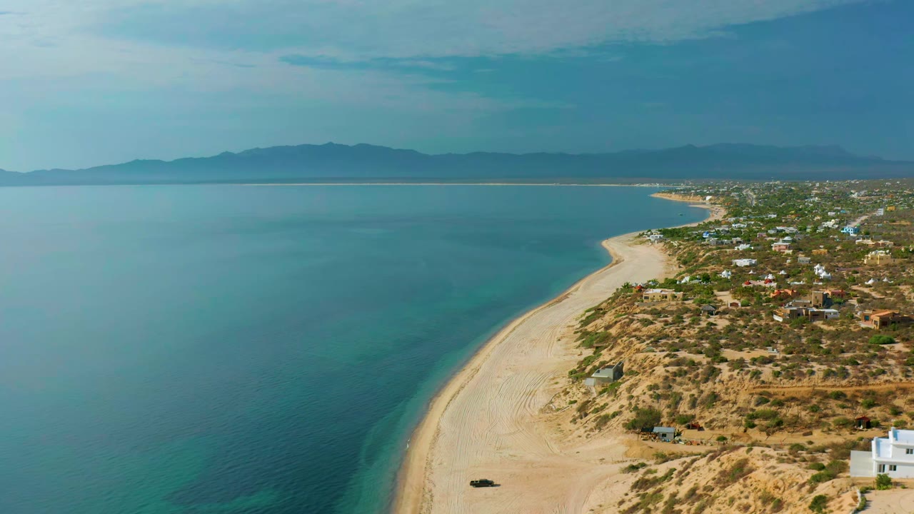 Aerial view over El Sargento La Ventana, Mexico, showing more open water to the left, the sandy shoreline running diagonally through the frame, coastal scrub and buildings to the right, and no visible people.