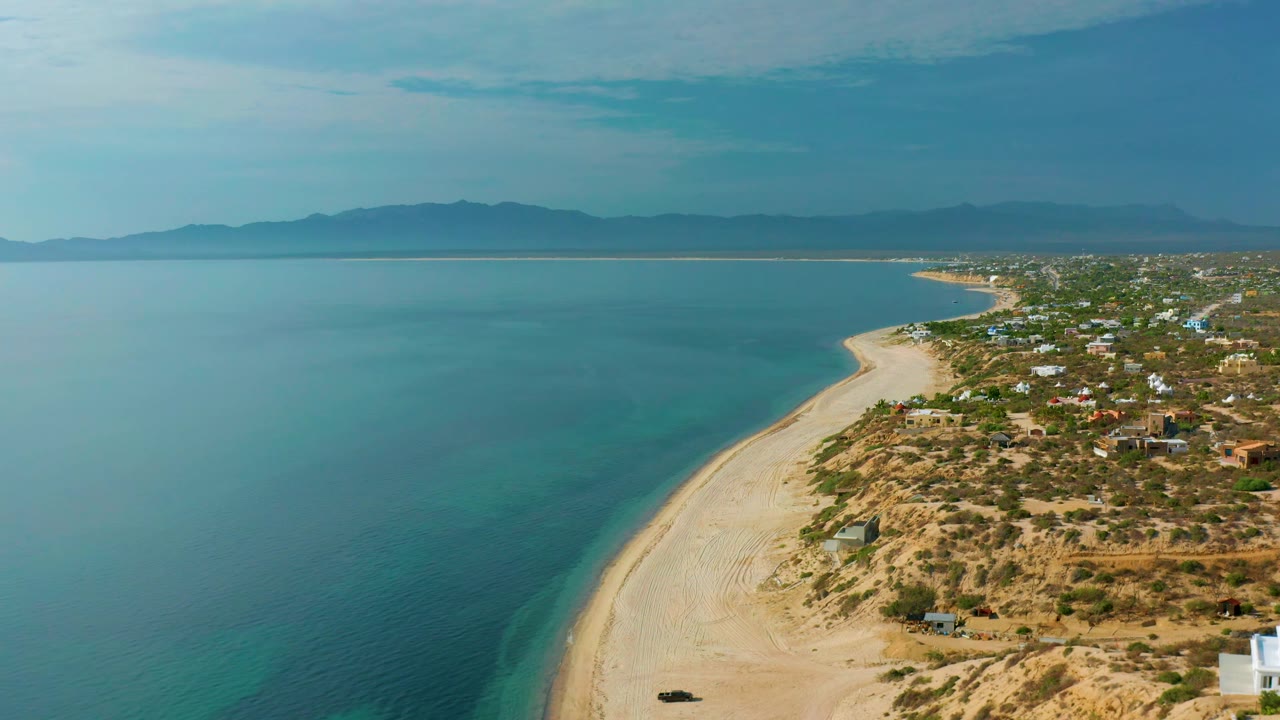 High aerial frame of El Sargento La Ventana, Mexico, with a clear-water edge tracing the sand, dry vegetation and low structures on the shore side, mountains across the bay, and no visible people.