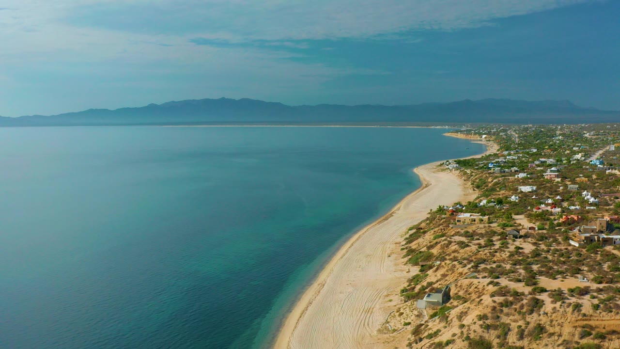 Opening aerial frame over El Sargento La Ventana with blue ocean on the left, a pale beach curve below, scattered shoreline buildings to the right, distant mountains, and no visible people.