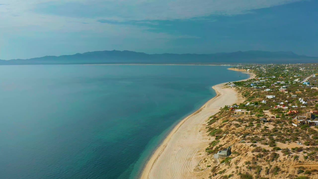 Aerial frame at 0:01 over El Sargento La Ventana showing more of the beach curve, calm turquoise water, low buildings near dry scrub, mountain horizon, and no visible people.