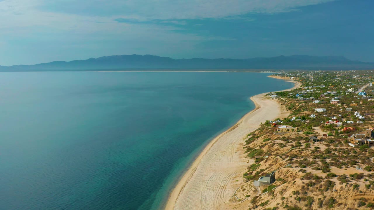 Frame at 0:02 shows El Sargento La Ventana from above with the sea taking most of the left side, the beach narrowing into the distance, scattered homes on the right, and no visible people.