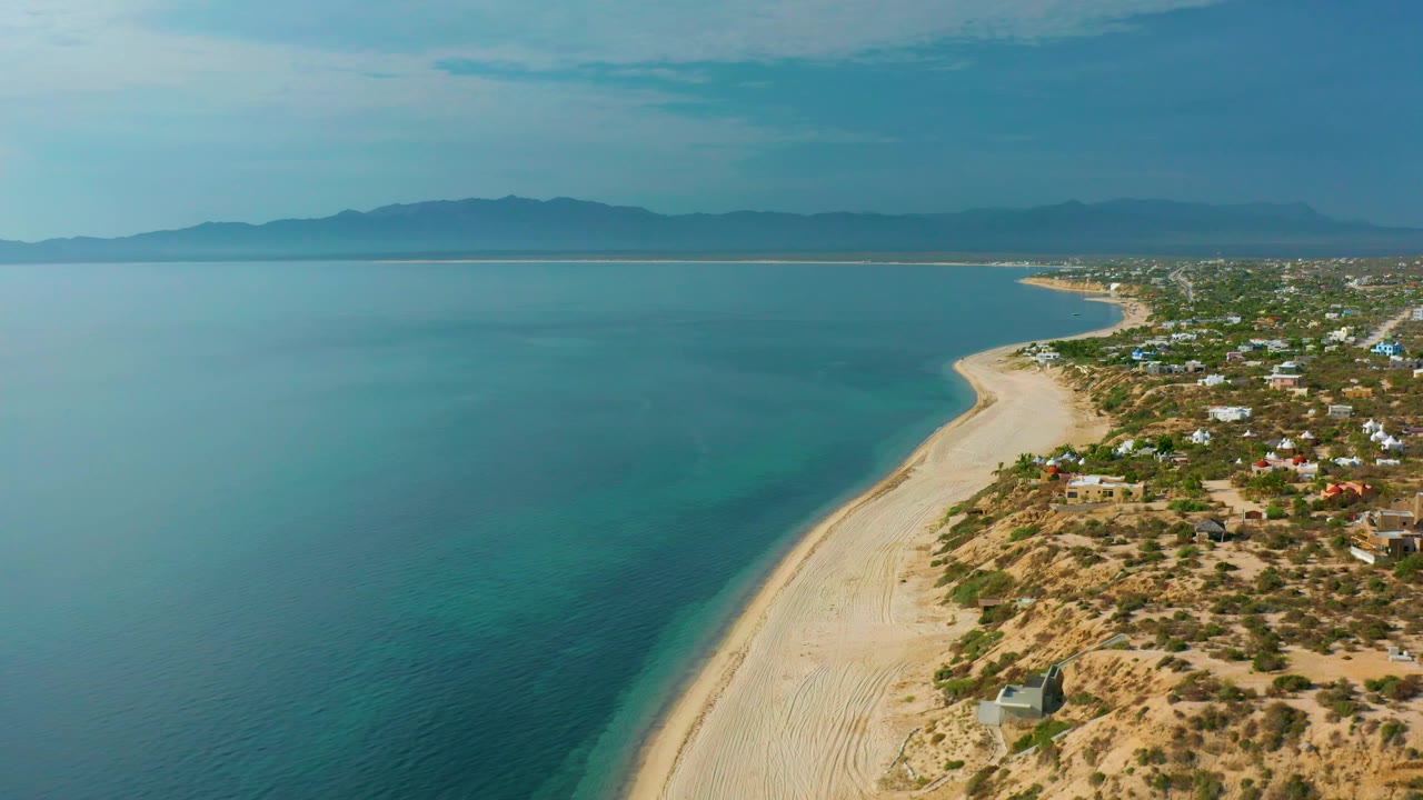 Aerial shoreline view with blue-green water, empty beach, desert scrub, and distant hills.
