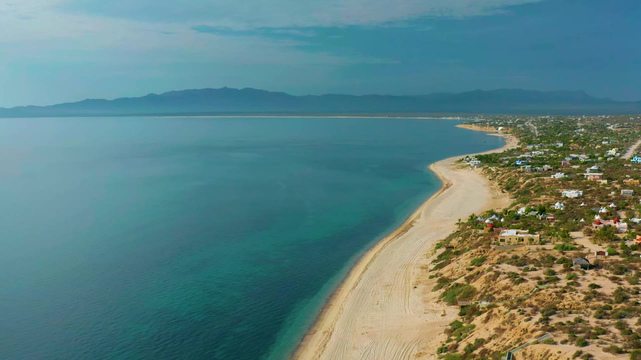 At 0:01, the drone view continues along the El Sargento La Ventana shoreline with turquoise shallows, pale sand, low village buildings, mountains, and no visible people.