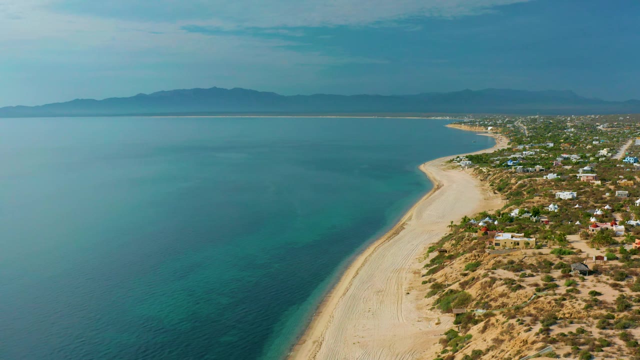 At 0:02, the aerial frame shows more of the curving El Sargento La Ventana beach, calm blue water to the left, dry lots and homes to the right, and no visible people.
