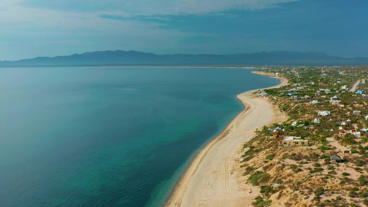 Aerial shoreline view with wide water, sandy beach, village, and mountain horizon.