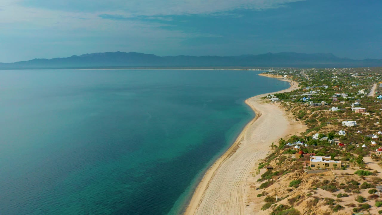 Opening aerial view over El Sargento La Ventana, Mexico, showing blue sea on the left, a pale beach curve, scattered coastal homes on dry land, distant mountains, and no visible people.