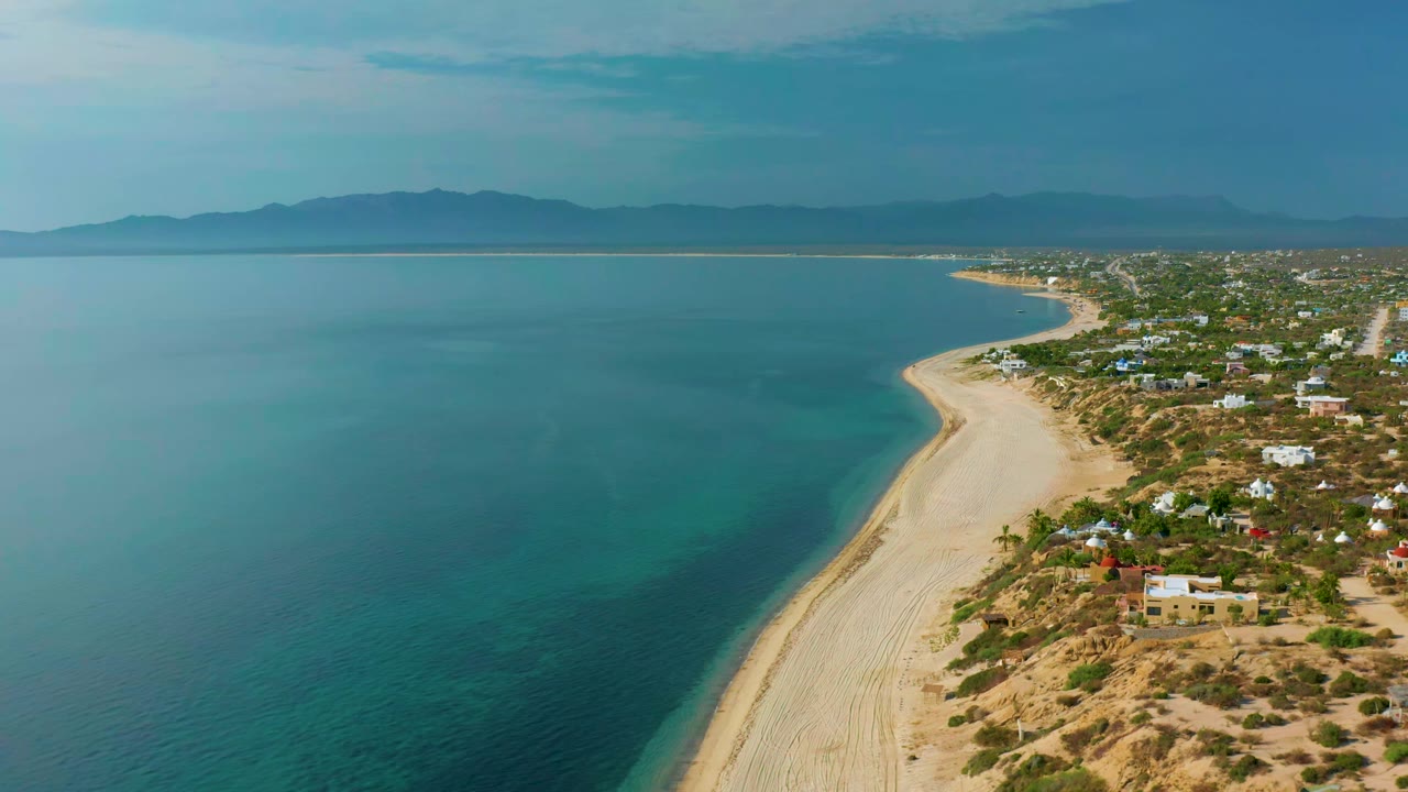 Aerial frame over El Sargento La Ventana with the shoreline curving deeper into the distance, blue water filling most of the left side, homes and scrubland on the right, mountains beyond, and no visible people.