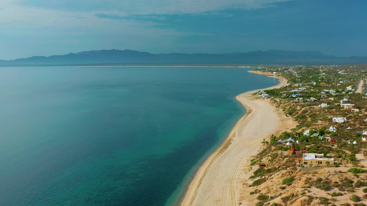 High view of El Sargento La Ventana coastline with a long sand strip between clear blue water and low homes, dry vegetation in the foreground, distant mountains, and no visible people.