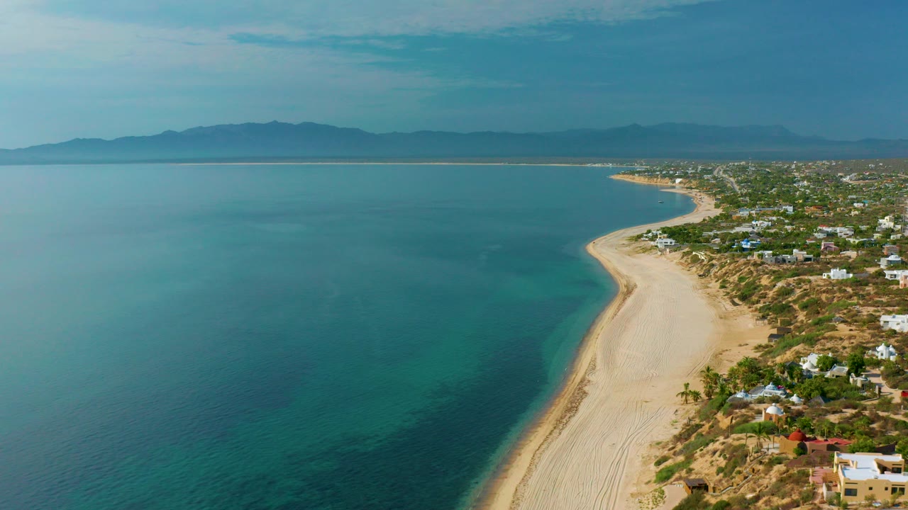 Opening aerial frame over El Sargento La Ventana, Mexico, showing a long curved sandy beach beside blue-green water, scattered coastal homes, hazy mountains, and no visible people.