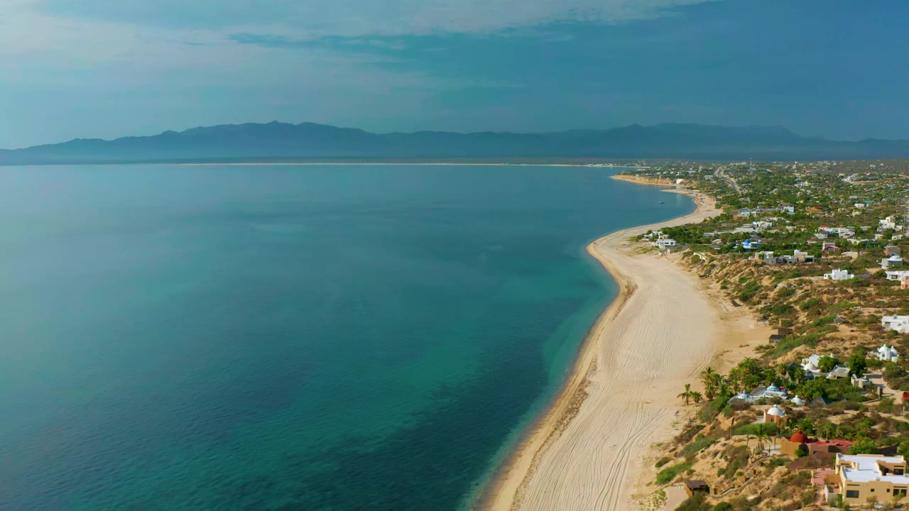 Wide aerial coastline with calm water on the left and houses along the sandy shore.
