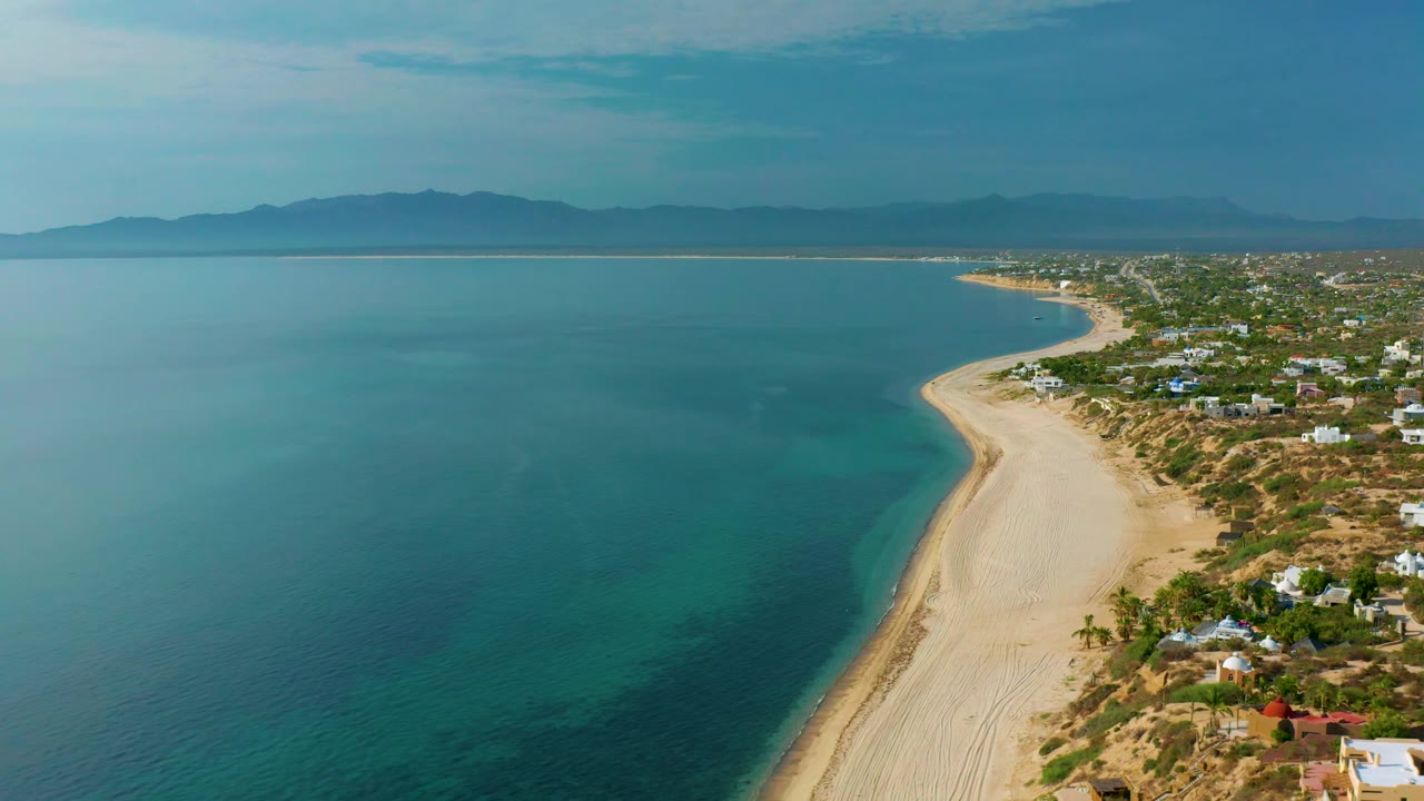 El Sargento La Ventana aerial view with turquoise shallows near the sand, darker water farther out, beach homes at frame right, distant mountains, and no visible people.