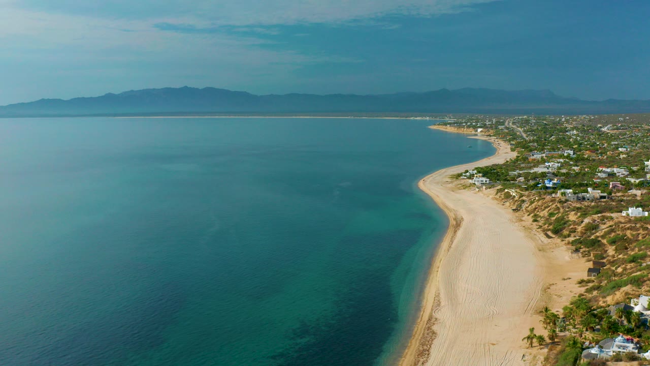High aerial frame over El Sargento La Ventana, Mexico, with blue sea on the left, a pale beach curving along the coast, low homes to the right, distant mountains, and no visible people.