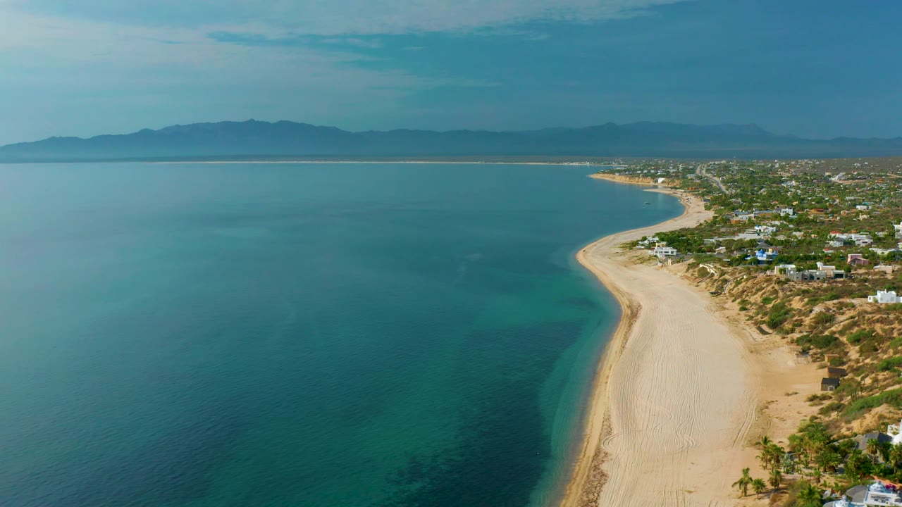 Wide aerial frame over El Sargento La Ventana, Mexico, showing calm blue sea filling the left side, a smooth sandy shoreline, scattered homes on the right, hazy mountains beyond, and no visible people.