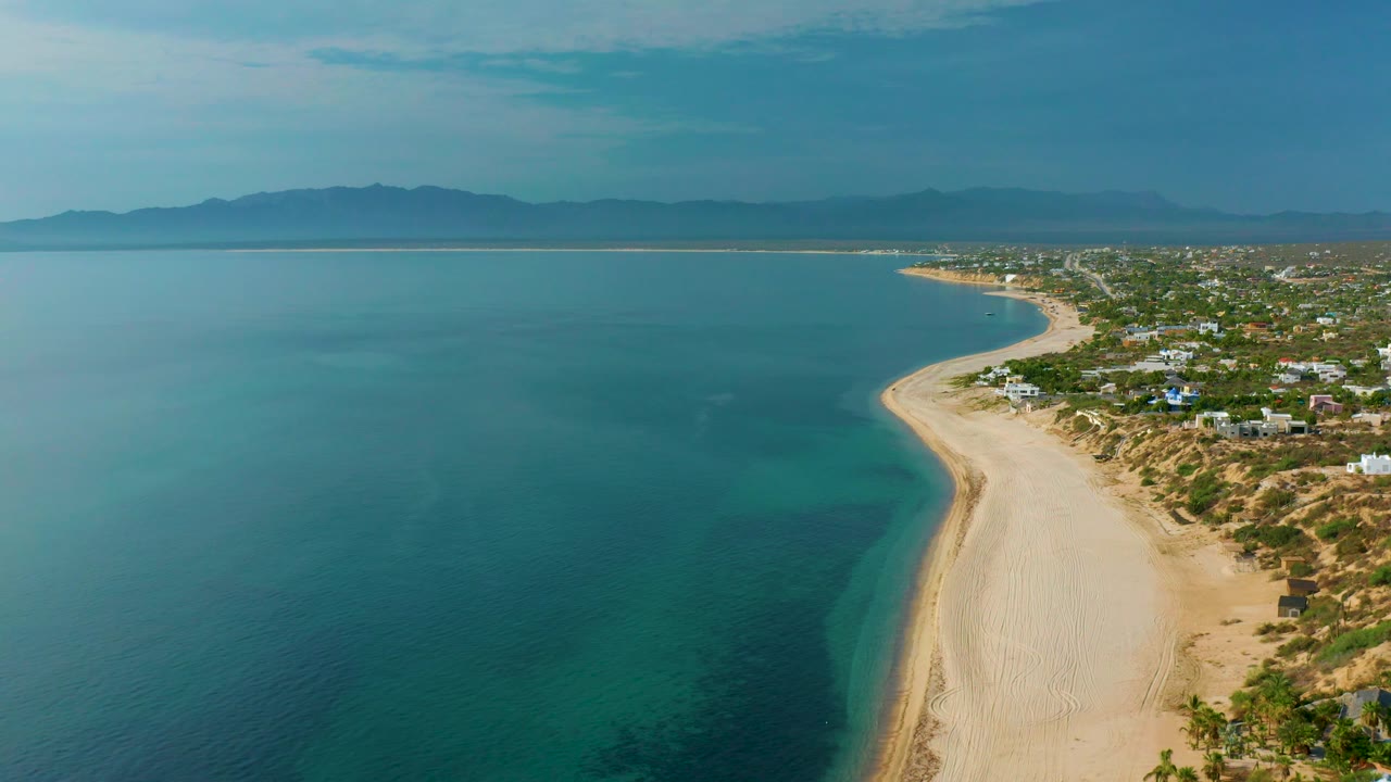 Blue ocean and broad sandy beach viewed from above with mountains in the distance.