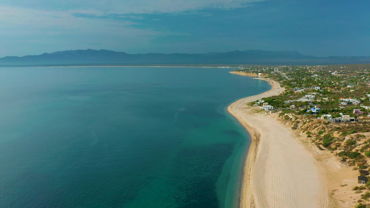 High aerial view over El Sargento La Ventana, Mexico, with blue-green water on the left, a wide sandy beach curving right, scattered coastal buildings, distant mountains, and no visible people.