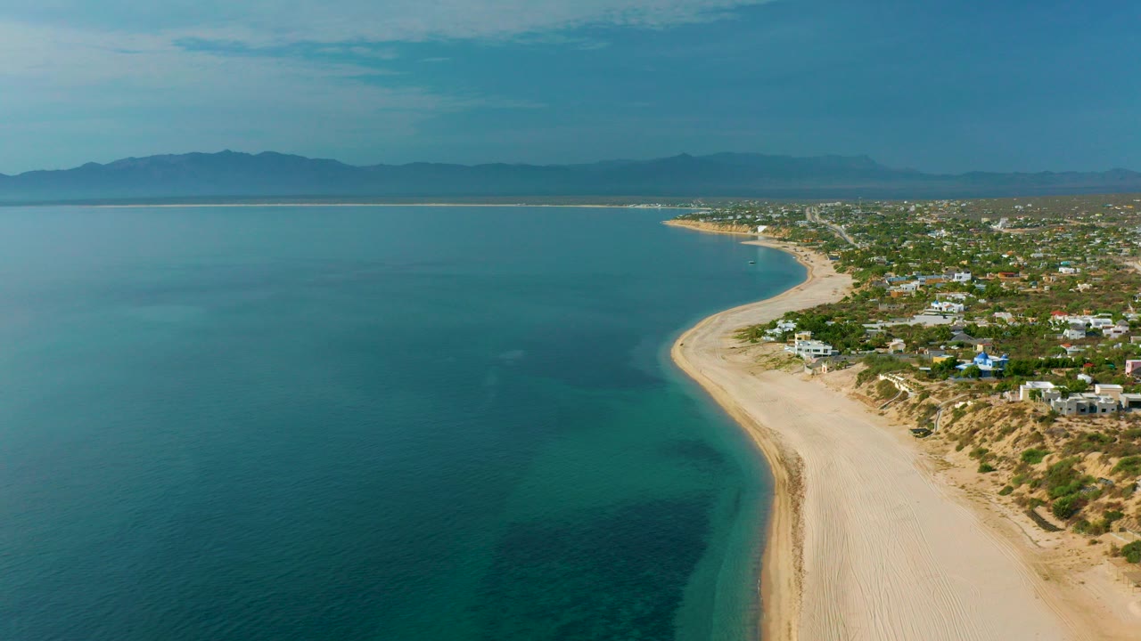 Opening aerial frame over El Sargento La Ventana, Mexico, with blue sea on the left, a pale sandy beach curving past shoreline homes, distant mountains, and no visible people.