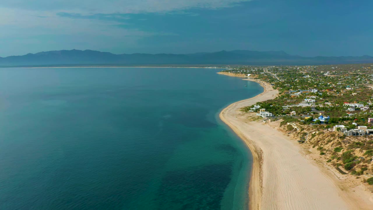 Wide aerial coastline with turquoise shallows, beach, homes, and hazy mountains.
