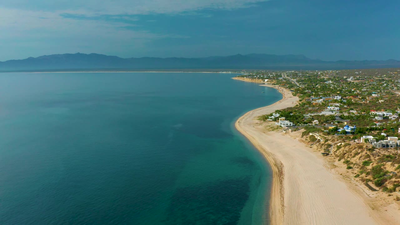 High aerial frame of El Sargento La Ventana with darker underwater patches in the sea, a pale beach running diagonally, homes and scrubland on the right, distant mountains, and no visible people.