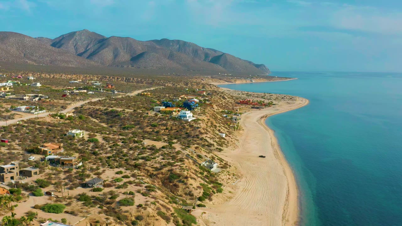Opening aerial frame over El Sargento La Ventana, Mexico, with scrubland and houses at left, a pale beach curving along turquoise water, mountains beyond, and no visible people.