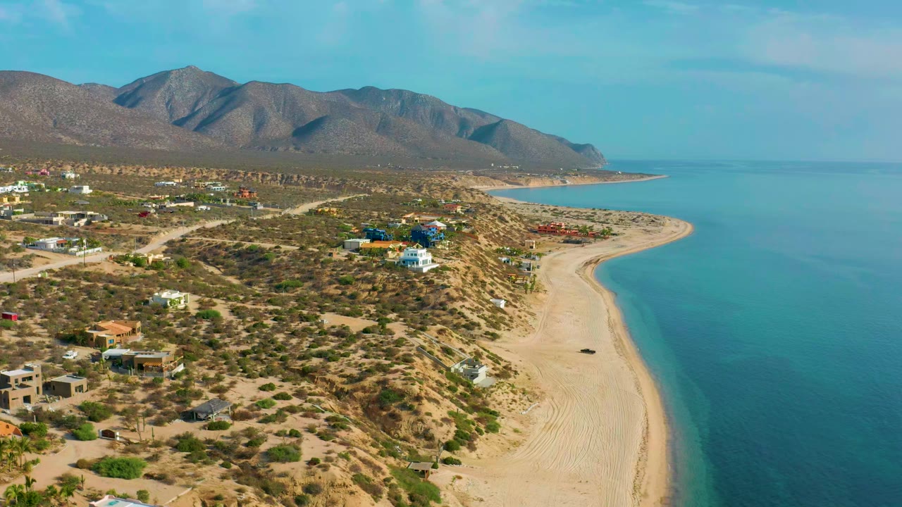 High coastal view with desert vegetation, beach curve, houses, mountains, and calm sea.